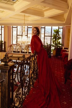 model in red long dress against the background of luxurious hotel interior. elegant young woman in long evening dress in hotel lobby with wrought iron railing. with evening make-up and hairstyle.