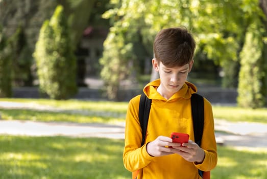 Teen boy in bright yellow hoodie looking at red smartphone screen while standing in front of green trees in daylight. Focus on modern youth lifestyle and digital connection.