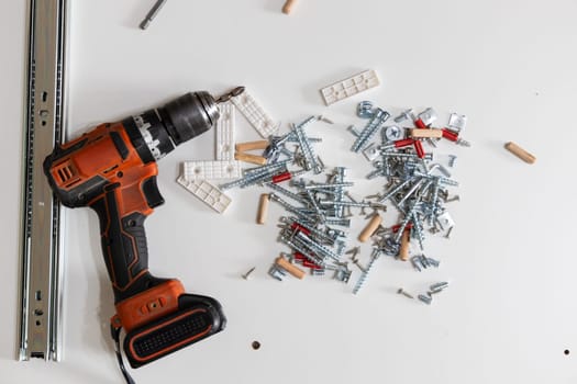 Electric screwdriver surrounded by various furniture fittings and hardware on clean white surface. Concept of home renovation, tools, and DIY assembly.