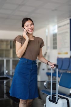 A young woman at the airport, smiling as she checks her phone while waiting for her flight, holding a suitcase and dressed casually.