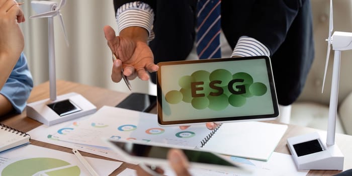 A professional presenting ESG data results on a tablet while discussing renewable energy technologies using wind turbine models during a meeting.