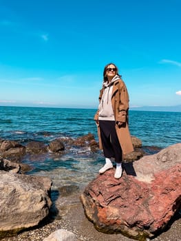 Young caucasian female standing on rocky shoreline in casual attire on sunny day.