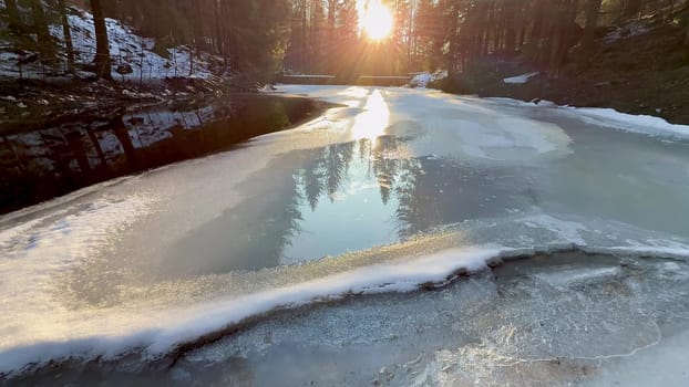 A tranquil scene with a partially frozen river reflecting the bright winter sun among tall, sturdy trees