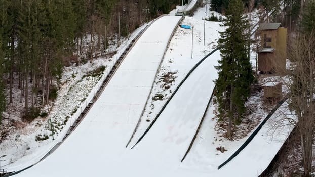 An aerial view shows a ski jumping ramp covered in fresh snow, surrounded by trees in a serene winter landscape