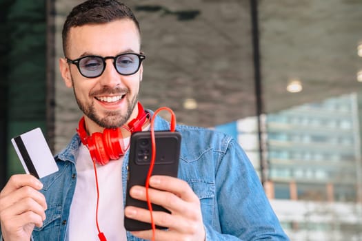 Confident man in casual outfit holds a blank business card and smartphone, smiling while listening to music through red earphones. Modern business and communication concept.