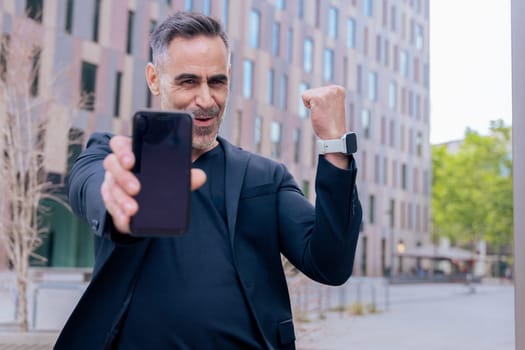 Happy man in black celebrates winning an online bet, holding up his smartphone and raising his fist in victory. He stands in a modern city area, full of joy and excitement.