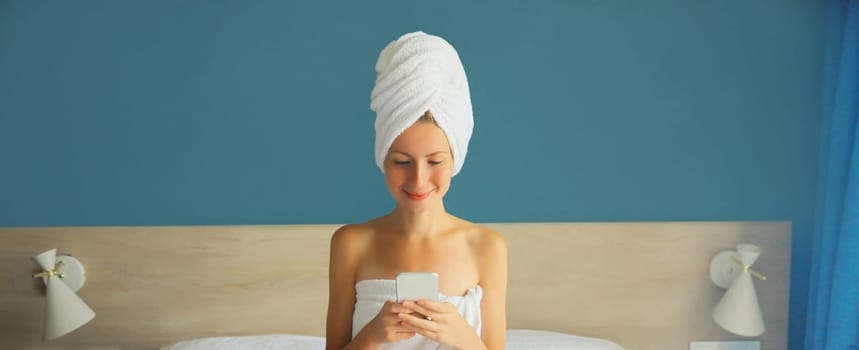Happy young woman looking at mobile phone drying her hair after bath with towel on her head sitting on white bed in bedroom in morning