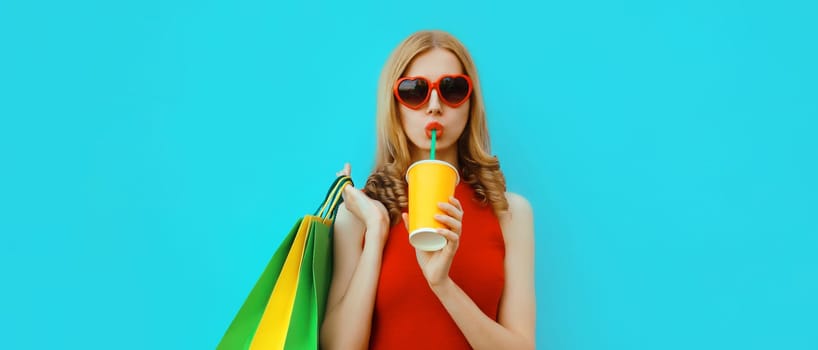 Portrait of beautiful relaxed young woman model with colorful shopping bags drinking juice in red heart shaped glasses on blue studio background