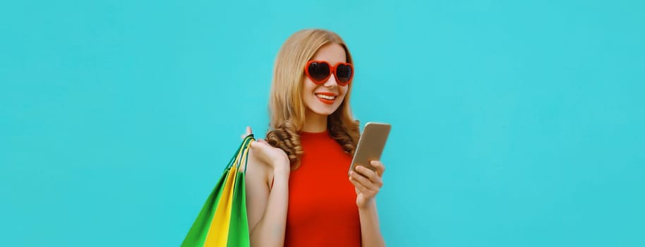 Beautiful happy young woman looking at smartphone with shopping bag in red dress with heart shaped sunglasses, girl holding phone on blue studio background