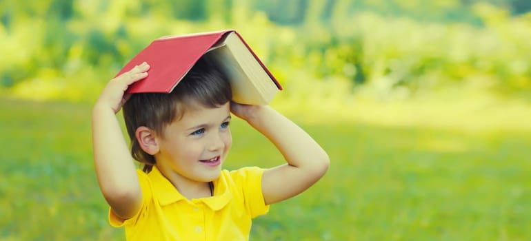 Happy little boy child reading a book sitting on green grass in summer park