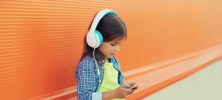 Portrait of little girl child with smartphone listening to music in blue wireless headphones, looking at device on orange background