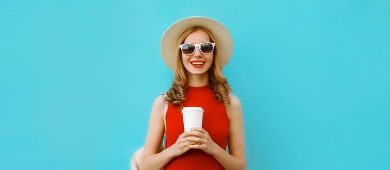 Portrait of beautiful happy young woman holding cup of coffee in summer tourist straw hat, red dress and glasses, pretty girl smiles with backpack on bright blue background