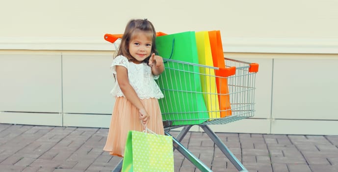 Happy little girl child with trolley cart and colorful shopping bags posing on city street