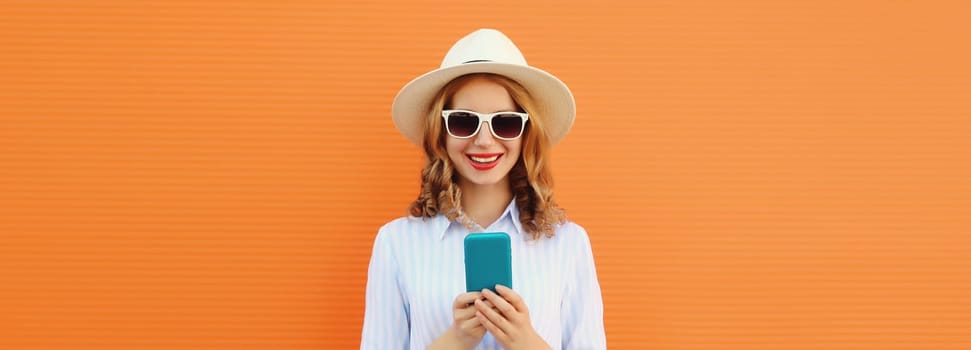 Summer portrait of happy traveler young woman with mobile phone, girl looking at smartphone wearing a straw tourist hat, casual clothing on orange studio background