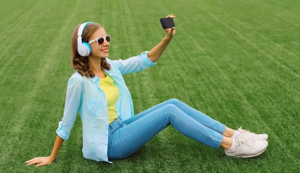 Happy young woman taking selfie with smartphone listening to music in headphones while lying on grass in summer park