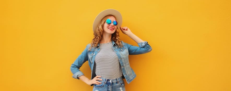 Stylish beautiful young woman wearing summer straw hat, jean jacket and shorts posing on orange wall studio background