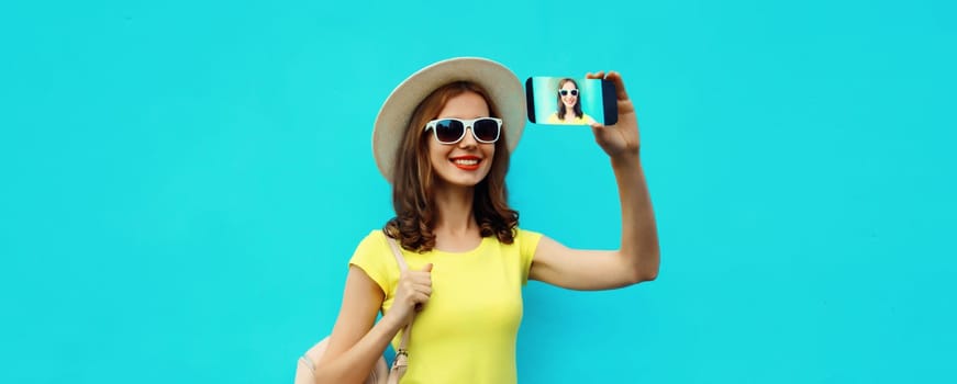Portrait of happy smiling young woman taking selfie with smartphone wearing a summer straw hat with backpack on blue background