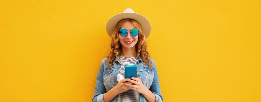 Summer portrait of happy traveler young woman with mobile phone looking at smartphone wearing a straw tourist hat, casual clothing on yellow studio background
