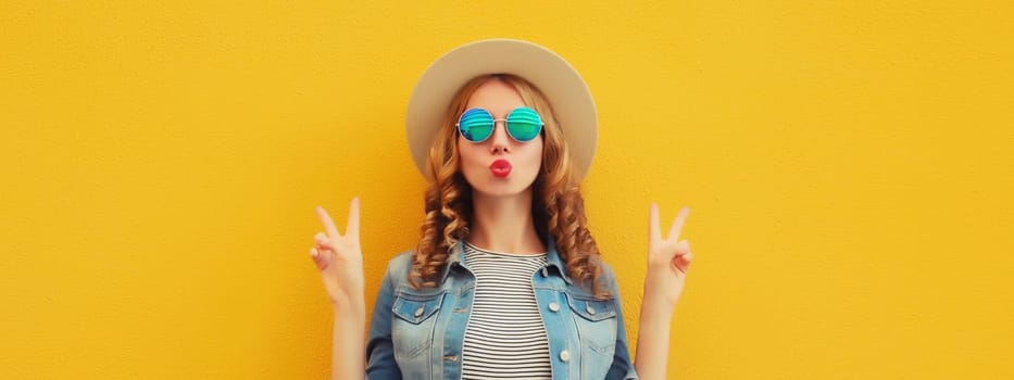Portrait of beautiful stylish young woman blowing kiss in summer hat, glasses, denim jacket posing on yellow studio background