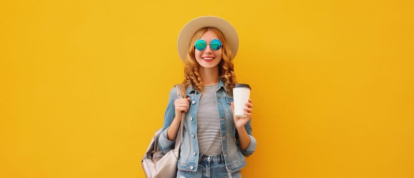 Beautiful stylish happy smiling young woman with cup of coffee wearing summer hat, backpack, jean jacket on bright wall yellow studio background
