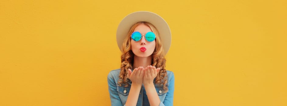 Portrait of beautiful stylish young woman blowing kiss in summer hat, glasses, denim jacket posing on yellow studio background
