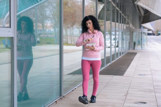 Middle adult Caucasian woman with curly black hair checking her smartwatch outdoors. She is wearing a pink sweatshirt and pink headphones, representing fitness, technology, and an active lifestyle.