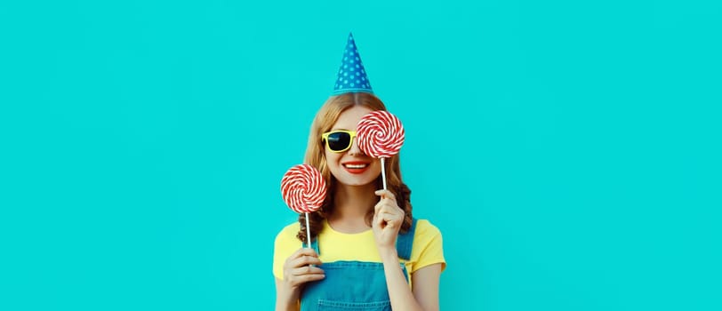 Holiday, celebration, event. Happy cheerful young woman laughs celebrating having fun at a party wearing festive birthday hat holding lollipop on blue background