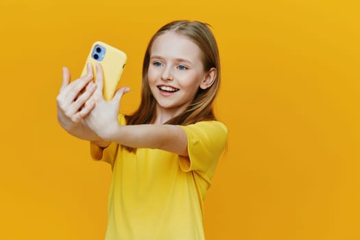A cheerful young girl captures a selfie with a yellow smartphone, expressing happiness against a vivid yellow backdrop, perfect for summer themes and social media concepts
