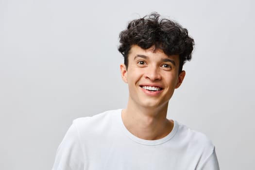 A cheerful young man with curly hair showcases a bright smile, set against a light gray background His white t shirt complements the minimalist style, perfect for conveying happiness and positivity