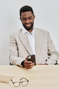 Confident man with dark hair and beard, wearing a beige blazer and white shirt, sitting at a desk and engaging with his mobile phone, in bright office space, relaxed and satisfied expression, indoor lifestyle scene, contemporary and professional atmosphere.