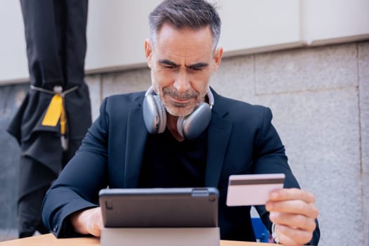 Confident caucasian man with headphones serious while using tablet and holding a credit card outdoors. Concept of online shopping, digital payment, modern lifestyle, and financial technology.