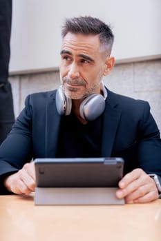 Focused caucasian businessman using a digital tablet at an outdoor cafe with headphones resting on his neck. Perfect for themes like remote work, technology, productivity,and modern lifestyle.Vertical