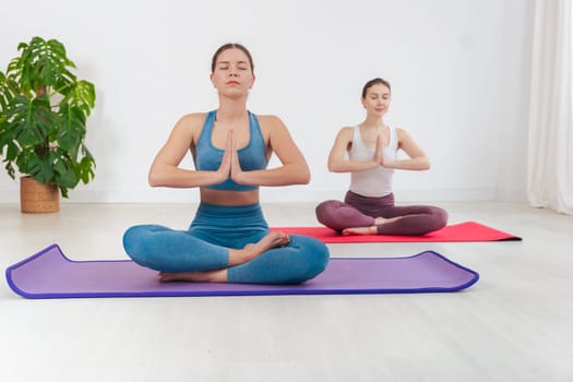 Two caucasian women doing yoga meditation indoors, sitting in lotus pose on mats. Calm and focused atmosphere in a bright minimal room, promoting mindfulness and physical wellness.