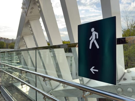 Close up of pedestrian walkway sign with walking figure and arrow on modern glass and steel bridge, with urban buildings and green trees visible in the background