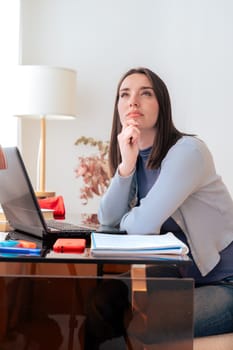 Caucasian woman sitting at desk, thinking while looking up. Laptop, notebook, and stationery on desk in modern home office. Creativity, concentration, decision-making, productivity, and contemplation.