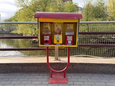 Colorful candy and gum vending machine stands on paved riverside path, inviting passersby to enjoy sweet treat. Surrounded by greenery, adds playful touch to the urban scene