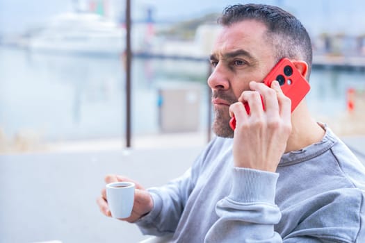 Man with short dark hair and beard drinks coffee while talking on his smartphone.He wears a casual gray sweatshirt and stands outdoors near the water, looking focused in a modern urban setting