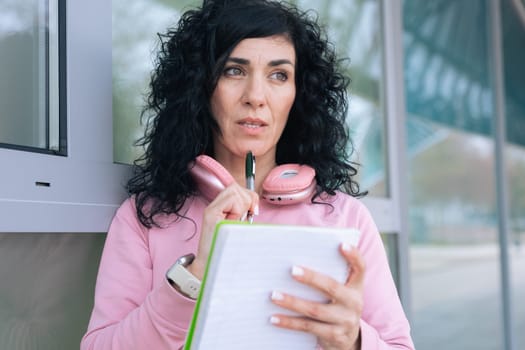 Caucasian sporty woman in a pink hoodie writing in a notebook outdoors. Wearing headphones around her neck, she appears focused and relaxed after a workout in an urban setting.