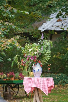 A bouquet of flowers in a vase on a table with a red and white checkered cloth, in the background a garden with trees and plants. Ideal for nature and garden lovers.