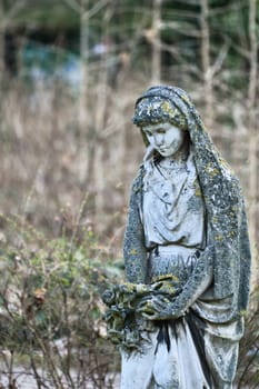 Moss-covered female statue with headscarf, surrounded by wild plants. The elegant sculpture holds flowers and stands in an enchanted garden. Mystical atmosphere.