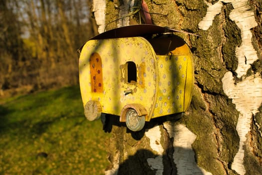 A yellow birdhouse in the shape of a caravan, attached to a tree trunk with white bark, in the sunlight. Ideal for nature and garden lovers.