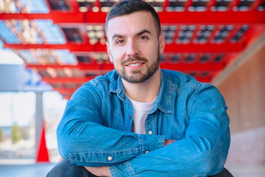 Young man with arms crossed looking directly at the camera under a red urban structure. Confident pose, casual style, and strong eye contact in a modern, outdoor city environment.