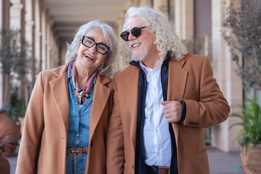 Smiling senior couple wearing sunglasses and brown jackets walking outdoors. They show elegance, joy, and companionship in a stylish urban environment with architectural background and plants.