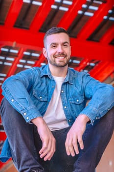 Vertical Portrait smiling young man sitting casually under a modern red urban structure. Relaxed pose, denim style, and confident expression in a bright. looking at camera