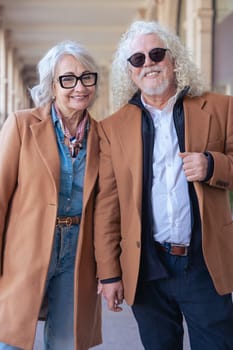 Vertical portrait of a smiling senior couple wearing sunglasses and stylish jackets. They walk outdoors confidently, showing elegance, happiness, and connection in an urban environment.