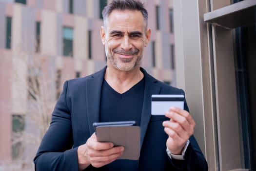 Confident caucasian man with headphones smiling while using tablet and holding a credit card outdoors. Concept of online shopping, digital payment, modern lifestyle, and financial technology.