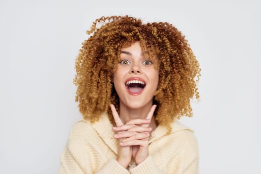 cheerful young woman with voluminous curly hair, showcasing a joyful expression on a plain light background She appears excited, with hands clasped and a bright smile, embodying positivity