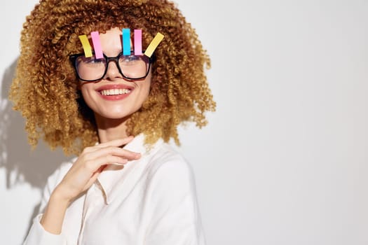 A cheerful young woman with voluminous curly hair and oversized glasses adorned with colorful clips smiles playfully against a simple white backdrop, showcasing a sense of creativity and joy