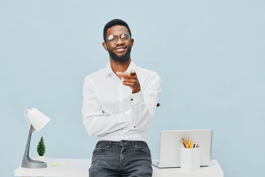 Cheerful confident man in casual office attire, wearing glasses and white shirt, standing at a desk with laptop, desk lamp, and potted plant in a bright, minimalistic workspace setting. Concept: professional success and positivity.