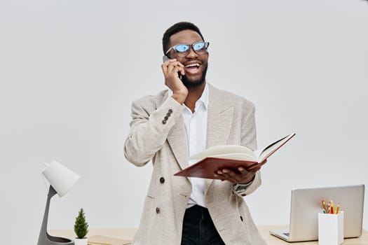 Smiling man with glasses using a smartphone and holding a notebook in a bright office setting with a minimalistic desk, laptop, and plant, displaying happiness and confidence, interior photography, professional business atmosphere, modern work environment. Concept: positive communication